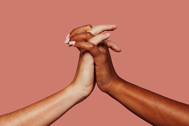 Feche o casal de mulheres multirraciais com mãos negras e caucasianas segurando um ao outro no pulso em amor de unidade de tolerância e conceito anti-racismo. Fundo rosa.